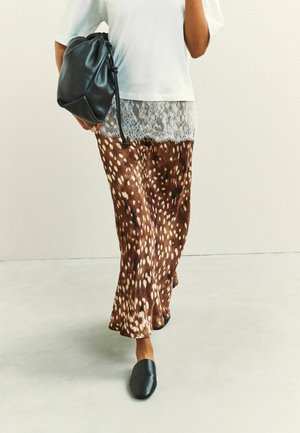 Person trägt braune und weiß gepunktete Hosen mit weitem Bein, weißes Oberteil mit Spitzenbesatz, schwarze Slipper und hält eine schwarze Ledertasche.