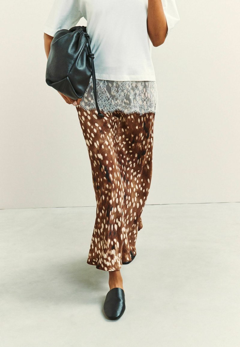 Personne portant un pantalon large marron et blanc à pois, un haut blanc orné de dentelle, des chaussures noires sans lacets, et tenant un sac en cuir noir.