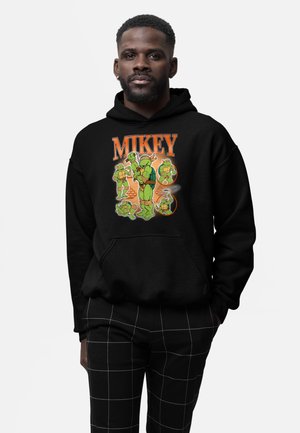 Hombre con sudadera con capucha negra con cinco personajes de las Tortugas Ninja Adolescente Mutantes y texto "MIKEY" en naranja encima de ellos.