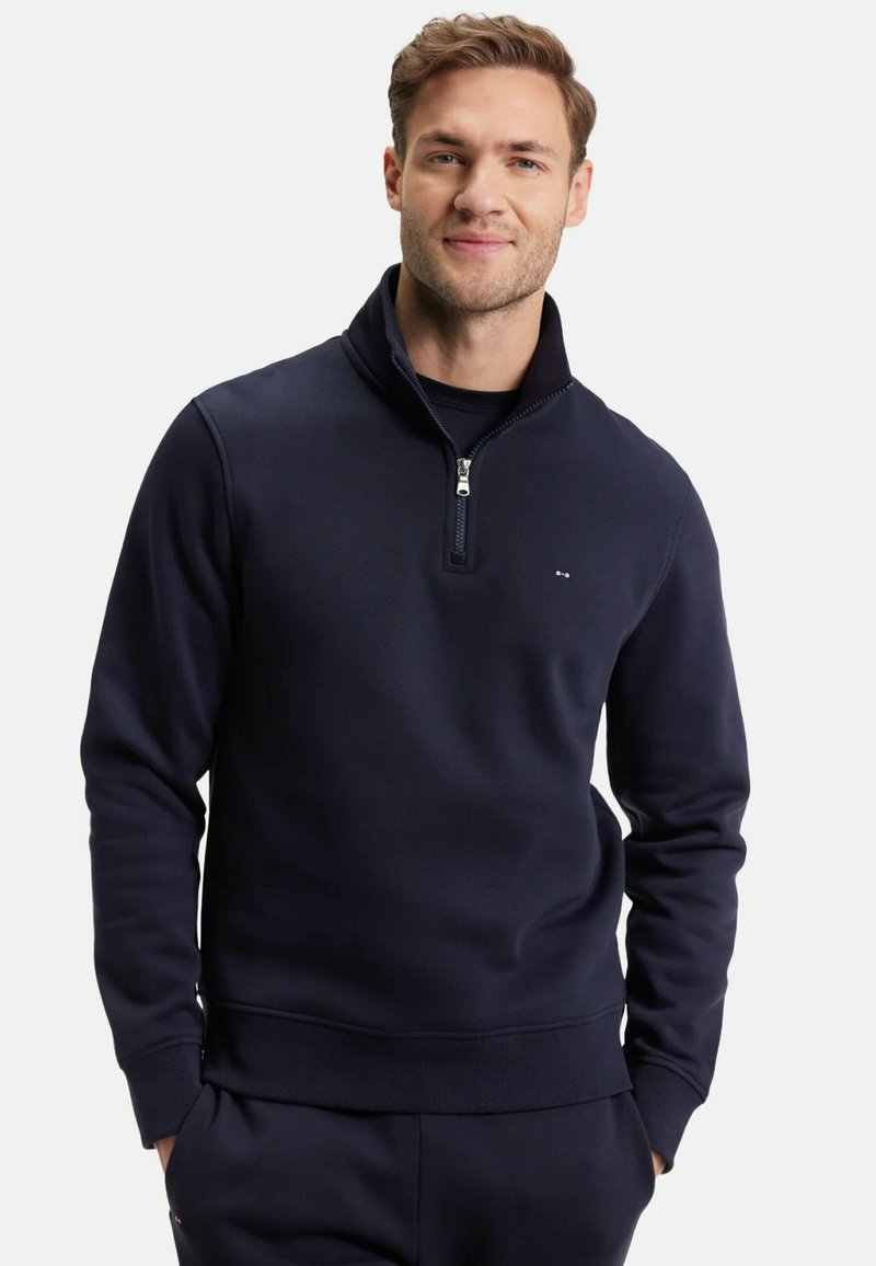 Sweatshirt zippé bleu marine en matériau doux, avec un col haut et des poignets côtelés, arborant un petit logo sur la partie gauche de la poitrine.