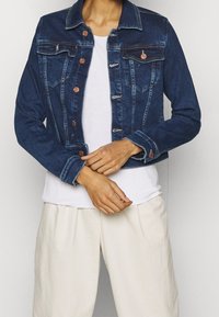 Denim jacket in dark blue with two chest pockets, copper buttons, and contrasting stitching. Worn over a white shirt and light-colored pants.