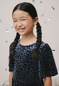 Tumši zila samta kleita ar melnu leoparda rakstu, īsām pūkainām piedurknēm un savāktu vidukli. Bērns ar pītām matu cirtām un spīduma akcentiem sejā.