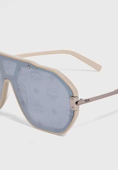 Lunettes de soleil à monture beige avec verres teintés bleus embossés du logo MCM et de formes de diamants, branches en métal gravées du nom de la marque MCM.