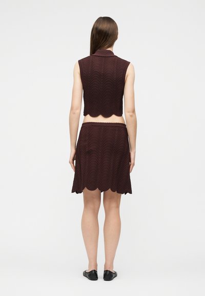 Femme portant un haut marron foncé sans manches à texture ondulée et une jupe assortie jusqu'aux genoux, debout de dos sur fond blanc.