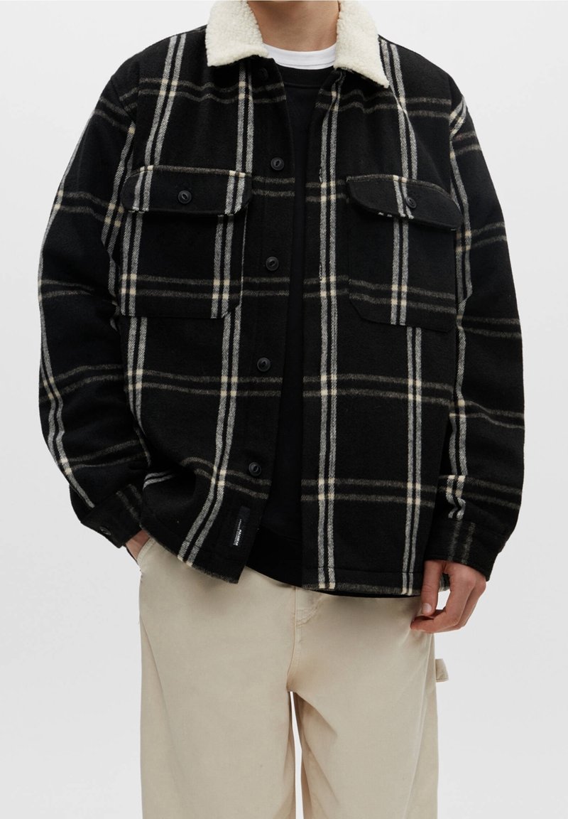 Uomo che indossa una giacca a quadri nera e beige con colletto in pelliccia bianca, camicia nera sotto e pantaloni beige, con le mani nelle tasche.