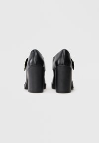 Sapatos de salto alto em couro preto, com bico arredondado e salto grosso. Acento de fivela prateada de um lado, textura suave, design minimalista.