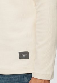 Crème-kleurig geribbeld shirt met lange mouwen en een zwart label. De stof heeft een gestructureerde uitstraling en sluit snugger aan rond de pols.
