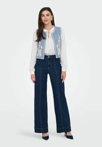 Ljusblå denimväst med blommig broderi, vit plisserad blus och högmidjade mörkblå vida jeans. Blå spetsiga klackar fulländar looken.