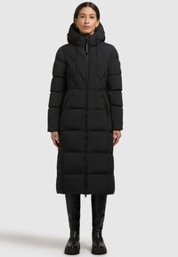 Abrigo acolchado negro con una silueta larga, capucha, cremallera frontal y dos bolsillos laterales. Presenta una textura acolchada y una construcción robusta.