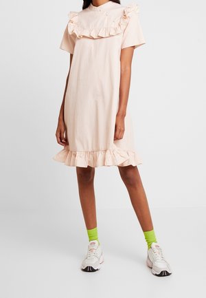 Jasnoróżowa sukienka w groszki, z krótkimi rękawami, marszczonym dekoltem i falbaną u dołu. Materiał wydaje się gładki, w połączeniu z białymi trampkami i zielonymi skarpetkami.