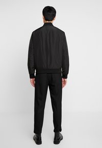 Veste bomber noire avec une texture lisse et brillante, poignets et ourlet côtelés, portée avec un pantalon noir et des bottes. Design simple et épuré.