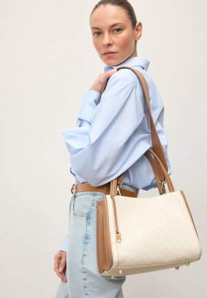 Beige håndtaske med lysebrune accenter og et tekstureret mønster, med guldfarvet hardware og dobbelte håndtag. Model iført en blå bluse og jeans.