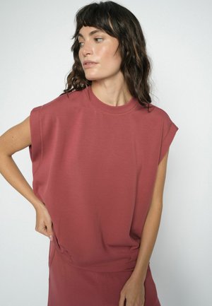 Žena s vlnitými tmavými vlasmi, ktorá má na sebe voľný bezrukávový vínový top, pozerá sa bokom s jednou rukou na páse na bielom pozadí.