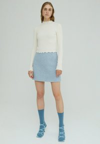 Cremfarbenes Langarmoberteil mit gewelltem Saum, kombiniert mit einem blauen Karorock im Mini-Format. Model trägt passende blaue Socken und Schuhe mit Kreuzriemen.