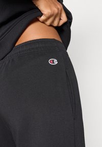 Μαύρο παντελόνι φόρμας με ελαστική μέση, πλαϊνές τσέπες και μικρό κεντημένο λογότυπο. Το ύφασμα φαίνεται μαλακό και με υφή.