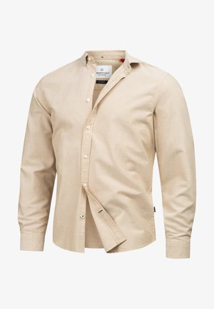 Beige nappapaita, joka on valmistettu pehmeästä puuvillasta, perinteisellä kauluksella, pitkillä hihoilla ja rintakehässä pienellä embleemillä. Malli on normaali.