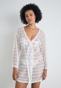 Cubierta de encaje blanca con diseño de escote en V, mangas largas, cintura con cordón y un colorido patrón floral debajo. Textura ligera y transpirable.