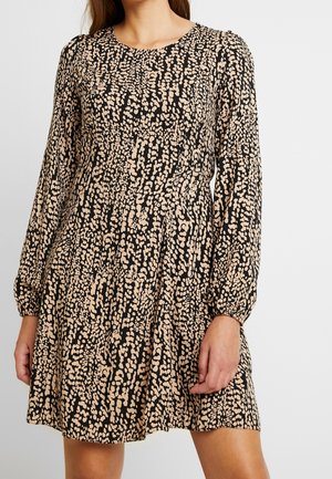 Femme portant une robe au-dessus du genou à manches longues, noire et beige à motifs tachetés, avec un col rond et une coupe ample.