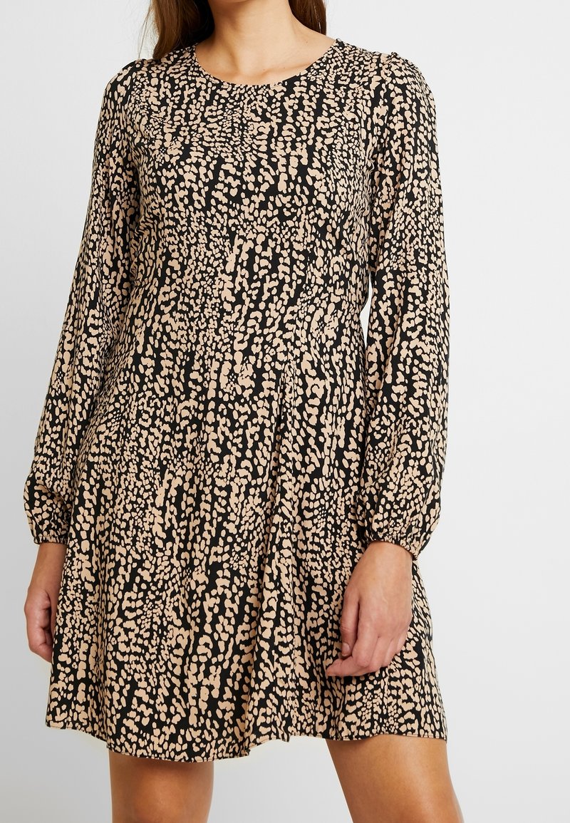 Femme portant une robe au-dessus du genou à manches longues, noire et beige à motifs tachetés, avec un col rond et une coupe ample.