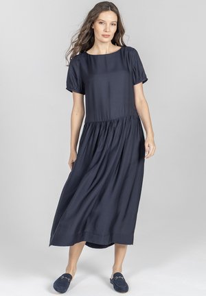 Kobieta stojąca, ubrana w granatową sukienkę midi z krótkim rękawem, z marszczoną talią oraz pasujące wsuwane mokasyny, na tle jednolitego szarego tła.