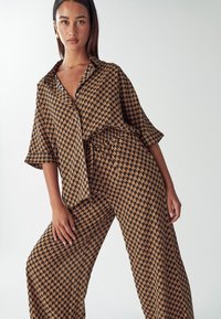 Smešani smeđi košulja s uzorkom i široke hlače koje imaju geometrijski dizajn. Tkanina ima glatku teksturu i labav kroj s kratkim rukavima.