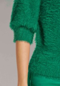 Grüner, flauschiger Pullover mit weichem Struktur, dreiviertellangen Ärmeln und engem Bund. Das Material wirkt samtig und gemütlich.