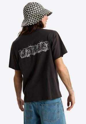 Osoba ubrana w czarną koszulkę z białą grafiką "VANS" na plecach, niebieskie jeansy i czarno-białą kraciastą czapkę bucket, zwrócona tyłem.