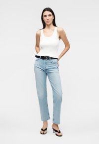 Mujer con una camiseta blanca sin mangas, vaqueros azul claro con cinturón negro y sandalias negras de tacón abierto, de pie contra un fondo blanco.
