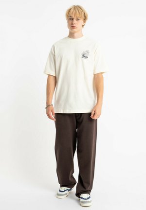 Jasnobeżowa bawełniana koszulka z krótkim rękawem i małym nadrukiem; zestawiona z luźnymi ciemnobrązowymi spodniami i białymi trampkami.