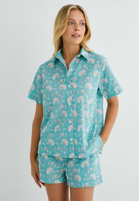 Conjunto de camisa de manga corta y pantalones cortos en turquesa con estampado floral blanco. Presenta un collar, cierre de botones en la parte delantera y bolsillos laterales.