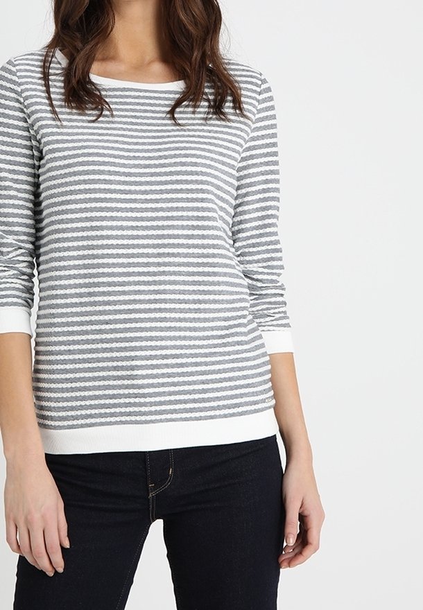 Femme portant un t-shirt à manches longues rayé horizontalement gris et blanc avec des poignets blancs et un pantalon foncé, debout devant un fond uni.