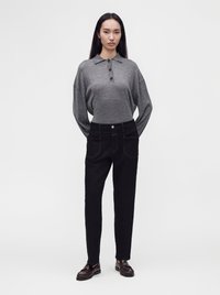 Jeune femme debout portant un polo gris à manches longues, un pantalon noir taille haute et des mocassins foncés devant un fond uni.
