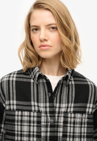 Black and gray plaid flannel shirt with buttons, collar, and chest pocket featuring logo detail. Soft fabric with a structured shape.