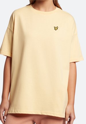 Person iført en løs, lysegul kortærmet t-shirt med en lille, broderet gul fuglelogo på venstre bryst.