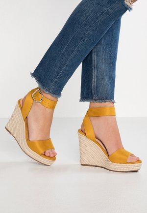Gule kile-sandaler med ankelremme brugt med blå jeans med frynset kant på hvid baggrund.