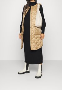 Beigevärinen pitkähihainen tikattu liivi, musta neulemekko, valkoiset nilkkurit mustilla yksityiskohdilla ja kermaan vivahtava olkalaukku. Sileä koostumus, rento design.