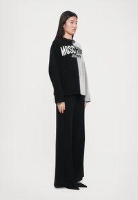Juodas ir pilkas megztinis su raštuotomis siūlėmis, kuriame yra baltas "Moschino Jeans" logo; derinamas su plačiomis juodomis kelnių.
