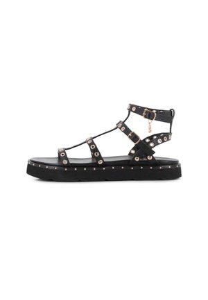 Sandalia plana negra con múltiples tiras, ojales dorados, hebillas y una suela gruesa con tachuelas decorativas.