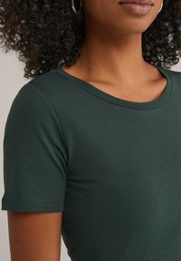 Close-up van een persoon met krullend haar die een donkergroen T-shirt met korte mouwen en ronde hals draagt, waarbij de nek en bovenkant van de torso zichtbaar zijn.
