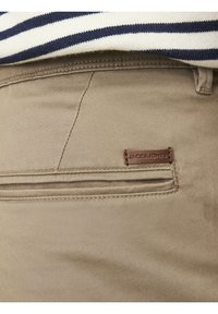 Primo piano di una tasca posteriore di pantaloni beige con un'etichetta cucita che recita "JACK&JONES" sotto una camicia a righe blu navy e bianca.