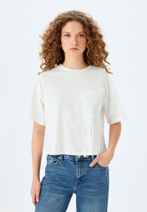 T-shirt en coton blanc à coupe courte, avec une poche en dentelle texturée et des détails en franges. Associé à un jean en denim bleu.
