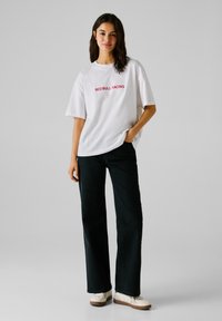 Kvinden bærer en hvid Red Bull Racing t-shirt, sorte brede jeans og hvide sneakers, mens hun står med den ene hånd i lommen.