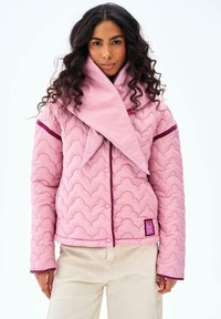 Žena s dlhými kučeravými vlasmi v svetloružovej prešívanej bunde s tmavoružovým lemovaním a veľkým skladaným golierom, v kombinácii s béžovými nohavicami.