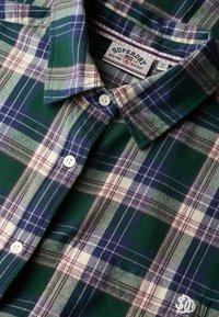 Chemise à carreaux vert, bleu, blanc et bordeaux avec des boutons blancs et un col visible, étiquetée "Superdry" à l'intérieur de l'encolure.