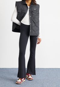 Svart vadderad väst med framfickor, tillsammans med figursydda svarta utsvängda byxor och flerfärgade sneakers med röda och vita detaljer.