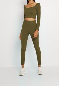 Top corto a coste verde oliva con maniche lunghe abbinato a leggings a vita alta coordinati. Il modello indossa scarpe da ginnastica bianche.