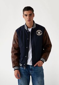 Chaqueta bomber de color azul marino con mangas de cuero marrón, cuello de canalé a rayas y parche con logo. Cuenta con botones a presión y cierre de cremallera. Ajuste casual.