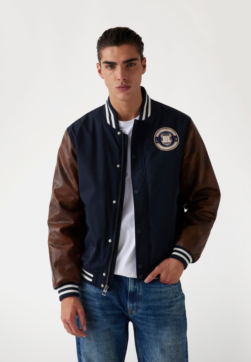 Chaqueta bomber de color azul marino con mangas de cuero marrón, cuello de canalé a rayas y parche con logo. Cuenta con botones a presión y cierre de cremallera. Ajuste casual.