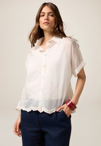 Blusa bianca a maniche corte con chiusura a bottoni, motivo floreale testurizzato, orlo a scallop, abbinata a pantaloni blu scuro e braccialetti rossi.