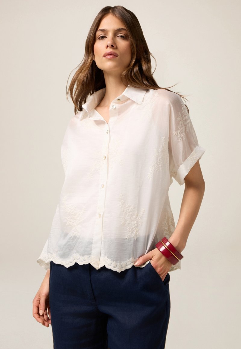 Blusa blanca de manga corta con botones en la parte delantera, patrón floral texturizado, dobladillo en forma de pico, combinada con pantalones azul oscuro y pulseras rojas.